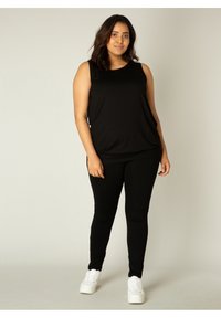 Zwarte mouwloze top gecombineerd met zwarte leggings, beide van een gladde stof. Het model draagt witte sneakers en staat voor een effen achtergrond.