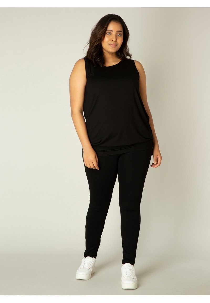 Zwarte mouwloze top gecombineerd met zwarte leggings, beide van een gladde stof. Het model draagt witte sneakers en staat voor een effen achtergrond.