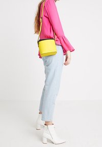 Sac bandoulière jaune de forme rectangulaire, avec une fermeture éclair noire et une sangle réglable. Porté avec un jean bleu clair et des bottines blanches.