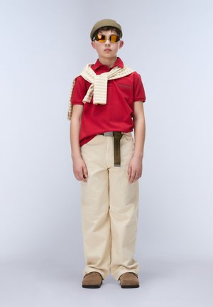 Garçon portant un polo rouge, un pantalon beige, des chaussures marron, un béret beige, des lunettes de soleil orange et un pull jaune rayé drapé sur les épaules, debout devant un fond uni.