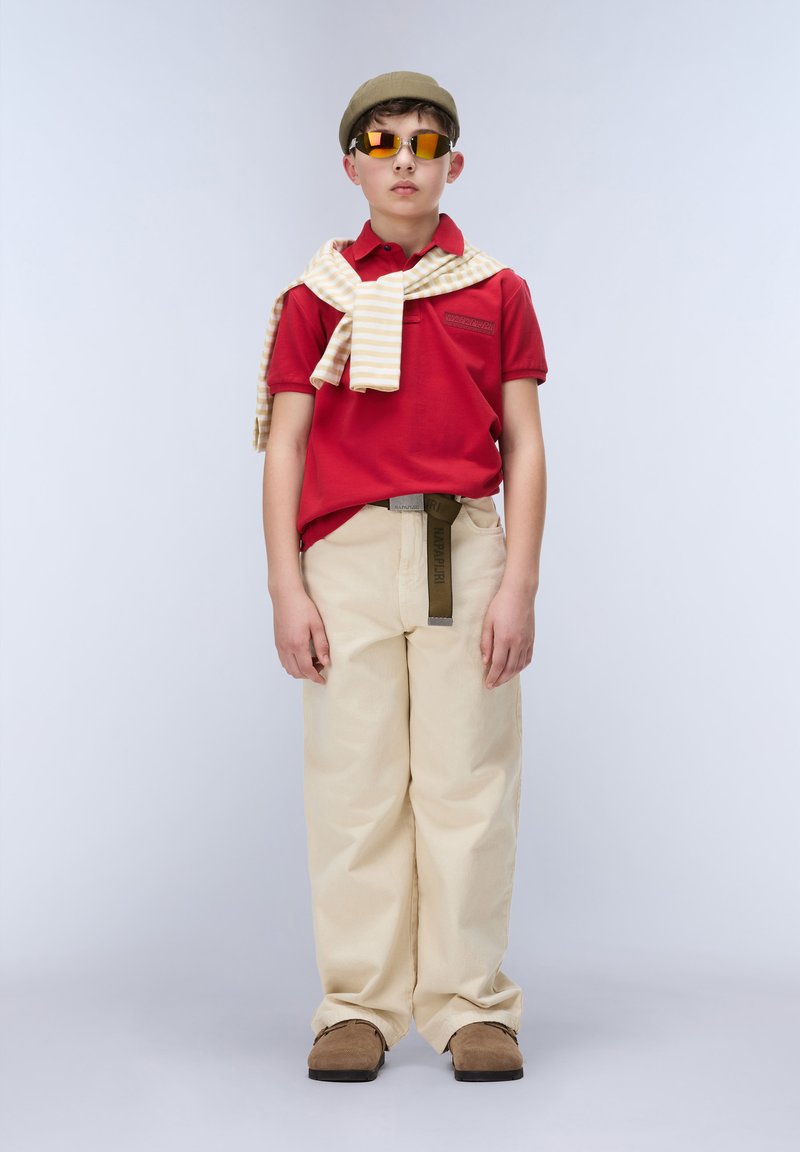 Garçon portant un polo rouge, un pantalon beige, des chaussures marron, un béret beige, des lunettes de soleil orange et un pull jaune rayé drapé sur les épaules, debout devant un fond uni.