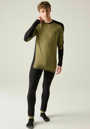 Young man adjusting olive and black long-sleeve athletic top, wearing matching black leggings and gray-toe socks, standing on neutral background.