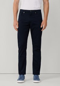 Pantalones oscuros de algodón suave, con un corte ajustado, bolsillos frontales y un cierre de botón en la cintura. Llevados con zapatillas de deporte en azul claro.