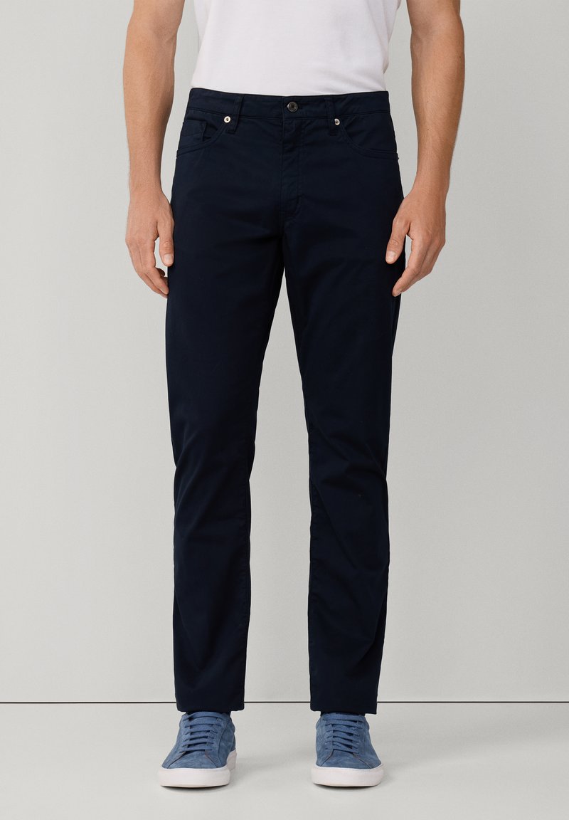 Pantalones oscuros de algodón suave, con un corte ajustado, bolsillos frontales y un cierre de botón en la cintura. Llevados con zapatillas de deporte en azul claro.