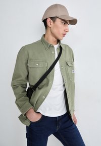 Olive green button-up shirt with two chest pockets, paired with a white t-shirt and dark blue jeans. Beige baseball cap and black crossbody bag.