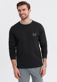 Ein Mann lächelt, trägt ein schwarzes Langarmhemd mit kleinem "EX"-Logo und grauen Nadelstreifenhosen, während er vor einem schlichten weißen Hintergrund steht.