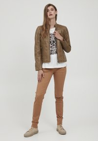 Hnedá kožená bunda so priliehavým strihom, spojená s bielym tričkom s grafickým motívom a svetlými nohavicami. Viditeľná je jemná textúra a zipsové vrecká.