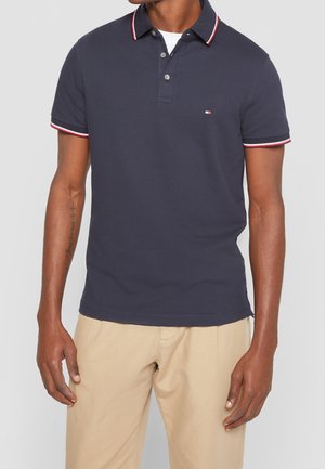 Homme portant un polo à manches courtes bleu marine avec une bordure blanche et rouge, pantalon beige, et un tatouage sur l'avant-bras gauche, debout devant un fond uni.