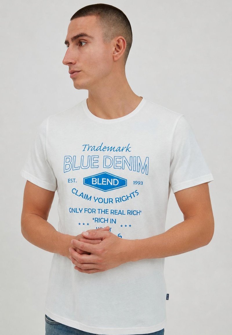 Homme aux cheveux courts portant un t-shirt blanc avec un texte bleu "Blue Denim Blend", debout devant un fond uni, mains jointes.