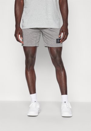 Miesmalli, joka pukeutunut harmaisiin teksturoituihin shortseihin, valkoisiin pitkävartisiin sukkiin, valkoisiin tennareihin ja vaaleanharmaaseen paitaan, seisoo valkoista taustaa vasten.