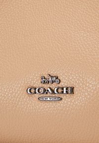 Surface en cuir beige texturé avec logo argenté de la marque Coach et emblème de calèche tirée par des chevaux au-dessus du texte « New York ».