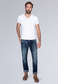 T-shirt bianca in cotone a V abbinata a jeans in denim blu scuro. Stivali in pelle marrone con lacci. Modello in piedi su uno sfondo neutro.