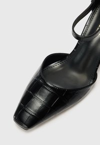 Schwarzer spitz zulaufender Schuh aus glattem, glänzendem Material mit einem Krokodilhaut-Muster, ausgestattet mit einem dünnen Knöchelriemen und einer offenen Ferse.