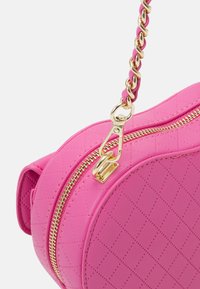 Kulatá růžová taška z umělé kůže s prošívanou texturou, zlatými zipy a řetízkovým popruhem. Stahovák zipu a akcentní kovové detaily ve zlaté barvě.