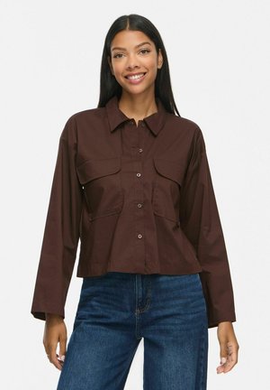 Femme aux longs cheveux noirs portant une chemise marron foncé boutonnée avec deux poches poitrine et un jean bleu, souriante sur fond blanc.