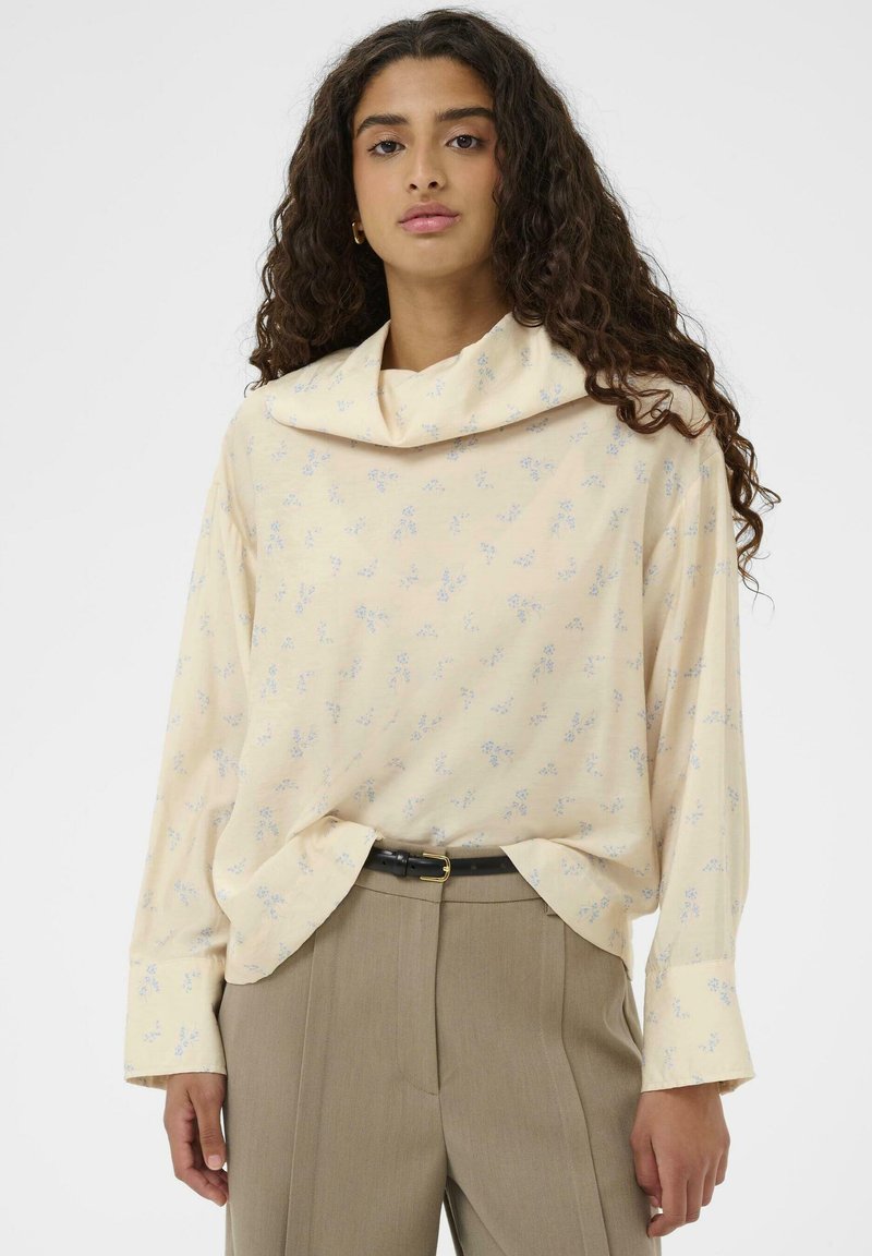 Ženska rumena cvetna bluza z dolgimi rokavi in padajočim izrezom, mehka tkanina in vzorci svetlo modrih cvetov. Skupaj z elegantnimi sivimi hlačami po meri.