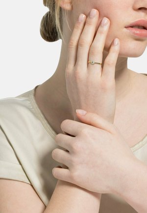 Goldring mit einem zentralen Edelstein, getragen an einer Hand mit neutralem Nagellack. Die Hand ruht an der Wange und zeigt eine glatte Hautstruktur.