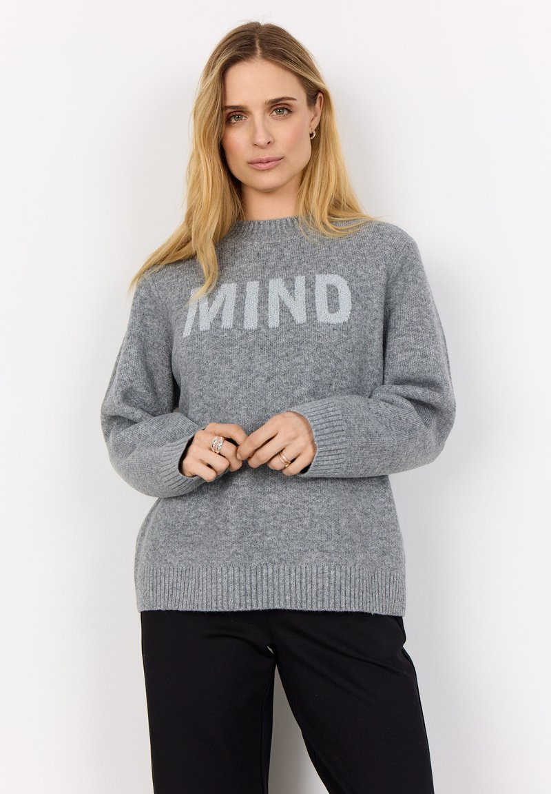 Grauer Strickpullover mit dem Schriftzug "MIND" auf der Vorderseite, Rundhalsausschnitt und gerippten Bündchen. Weiche Textur mit lässiger Passform. Kombiniert mit schwarzer Hose.