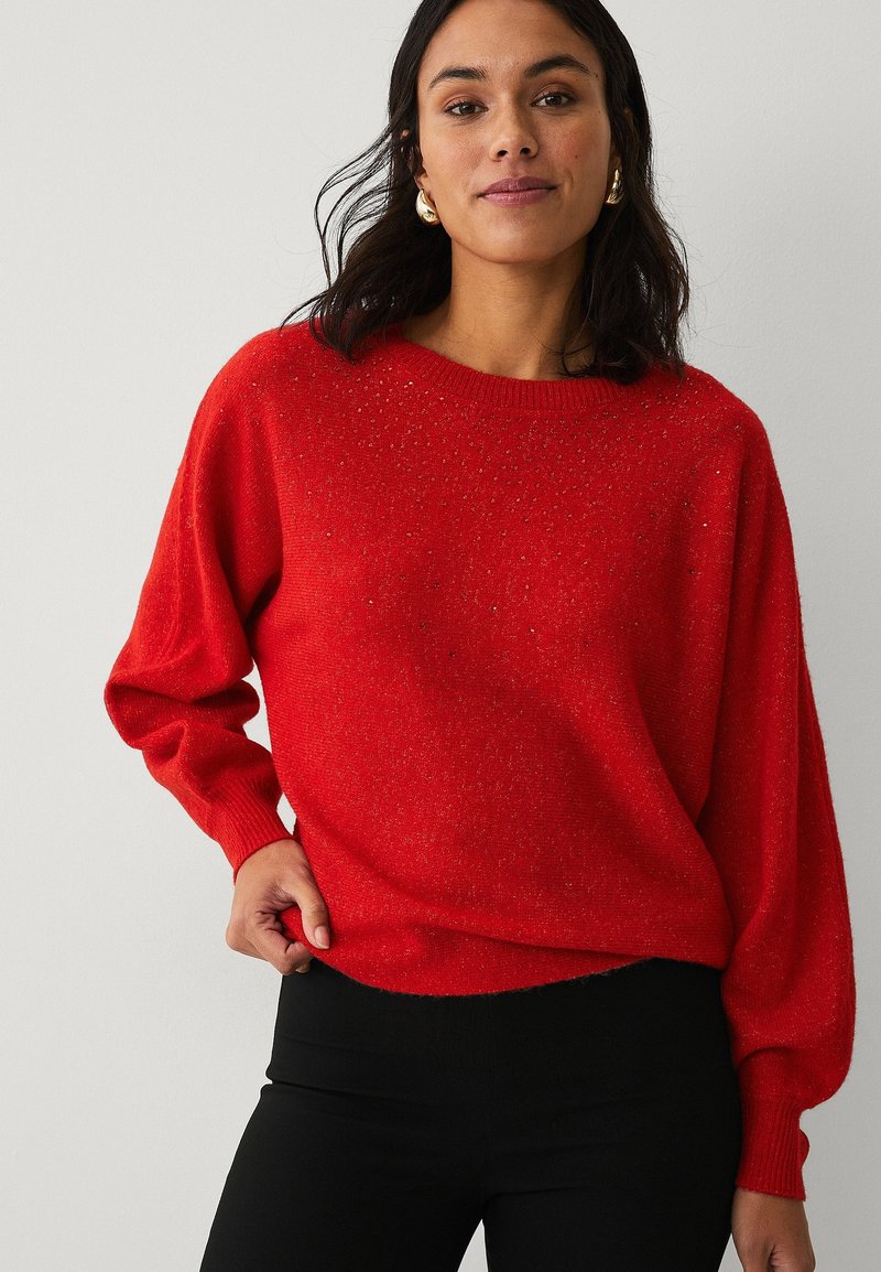 Rød, løstsiddende sweater med rund halsudskæring og lange, puffede ærmer. Det strukturerede stof har subtile glitrende detaljer. Sammen med sorte underdele.