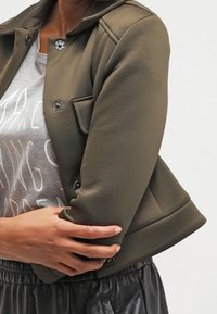 Persona vistiendo una chaqueta de botón verde oliva sobre una camiseta gráfica gris y pantalones de cuero negros, con los brazos cruzados a la altura de la cintura.