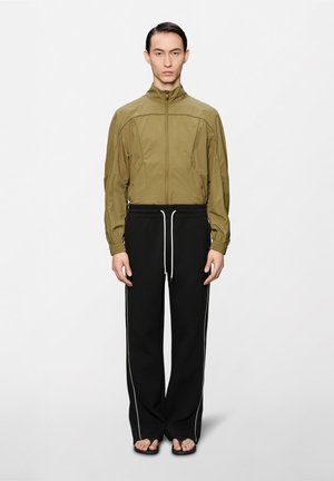 Vékony férfi olívazöld cipzáras dzsekiben, fekete, fehér oldalsávos bő szárú nadrágban és fekete papucsban, sima fehér háttér előtt állva.