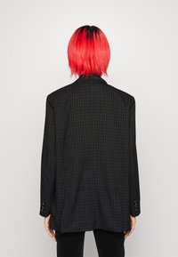 Černý kostkovaný blejzr s volným střihem, zdobený modrými mřížkami a knoflíkovými manžetami, zobrazený zezadu na neutrálním pozadí.