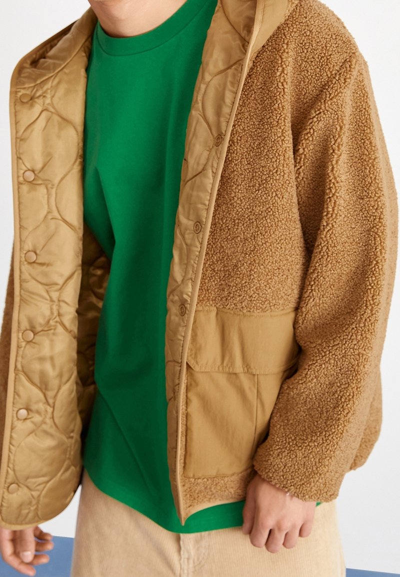 Chaqueta de forro polar marrón con un revestimiento acolchado beige, que presenta un bolsillo frontal y botones. Llevada sobre una camiseta verde. Textura suave, diseño casual.