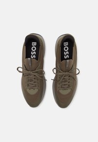 Sapatilhas verde-oliva com parte superior em malha, bico redondo, sola de borracha texturizada e atacadores planos. Apresenta a marca "BOSS" na língua e na palmilha.