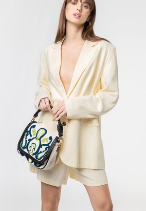 Donna che indossa un blazer crema oversize e pantaloncini, che tiene in mano una borsa beige con motivo ricamato blu, verde e nero.