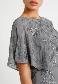 Kobieta ubrana w szaro-suknię z krótkimi rękawami, ozdobioną skomplikowanym haftem kwiatowym z ciemnych koralików i cekinów.