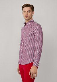Camisa de botones a rayas en rojo y blanco, con cuello extendido, un bolsillo en el pecho y mangas largas con puños abotonados. Llevada con pantalones rojos.