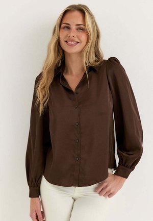 Leende kvinna med långt blont hår, klädd i en mörkbrun knäppt blus och vita byxor, stående mot en enkel ljus bakgrund.