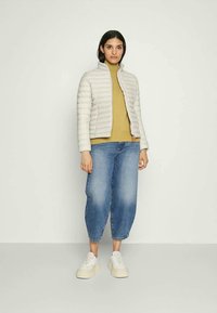 Mulher em pé, vestindo uma jaqueta puffer bege clara, um suéter de gola alta mostarda, jeans azuis largos e ténis brancos de plataforma, em um fundo simples.