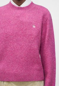 Personne portant un pull en maille texturée rose sur une chemise blanche à col, avec un pantalon beige visible en bas.