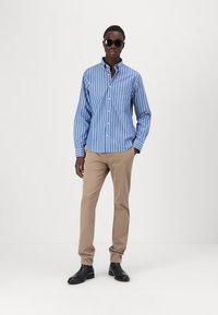 Lange mouwen, lichtblauw en wit gestreept shirt met een zak, gecombineerd met beige broeken en zwarte schoenen, aangevuld met een zonnebril in het zwart.