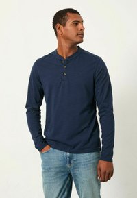 Marineblaues Lange-Knopf-Henley-Shirt mit drei Knöpfen, geripptem Kragen und dezentem Logo. Getragen mit hellblauen Jeans in entspannter Passform.