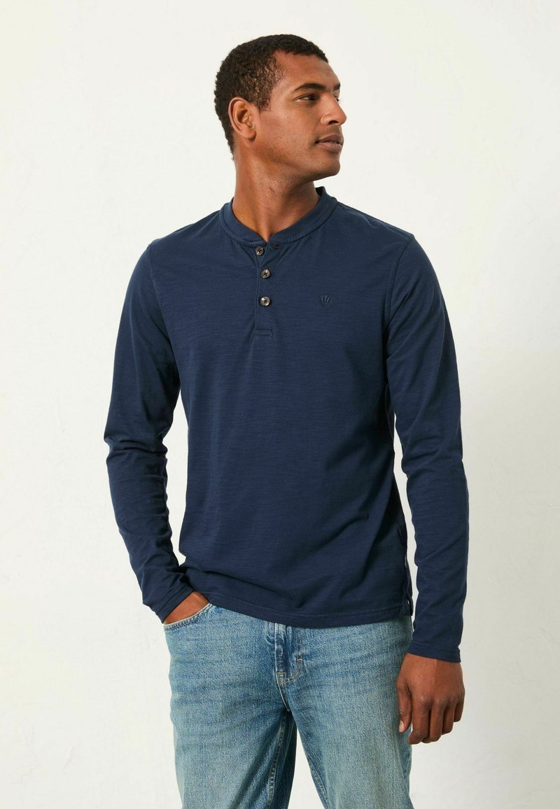 Marineblaues Lange-Knopf-Henley-Shirt mit drei Knöpfen, geripptem Kragen und dezentem Logo. Getragen mit hellblauen Jeans in entspannter Passform.
