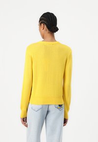 Pull jaune à manches longues avec un col rond et un ourlet côtelé. Porté avec un jean bleu clair présentant un patch de dessin animé sur la poche arrière.