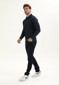 Marineblauwe polo met lange mouwen en een kraag, voorzien van een subtiele logo. Gepaard met donkere jeans en witte sneakers met zwarte accenten.