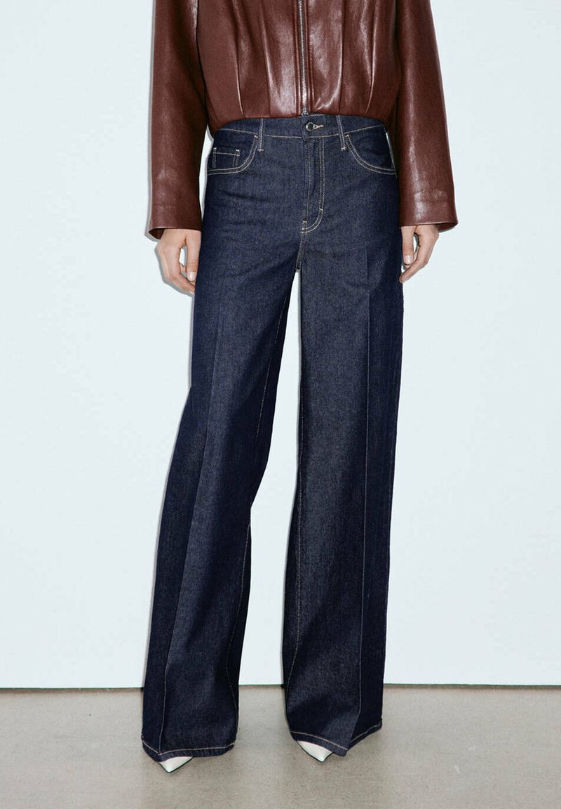 Personne portant un jean large foncé et une veste zippée en cuir marron, se tenant sur un sol et un mur unis de couleur claire.