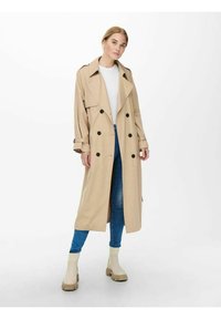 Beige trenchcoat med dobbeltknapper, bred krage og belte i ermene. Brukt over en hvit topp og blå jeans, kombinert med beige støvler.