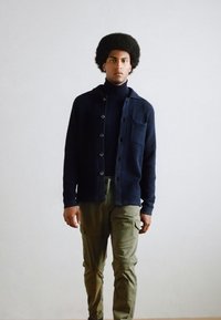 Mann mit Afro-Frisur, der eine schwarze Knopfpulloverjacke, einen schwarzen Rollkragenpullover und olivgrüne Cargo-Hosen trägt und vor einer neutralen hellen Wand steht.