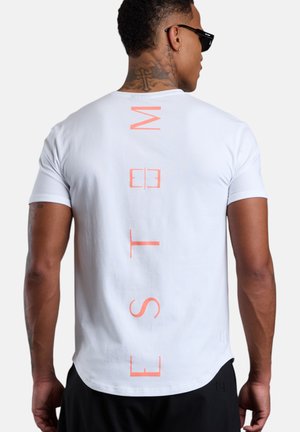 Mann trägt weißes Kurzarm-T-Shirt mit vertikalen pfirsichfarbenen Buchstaben "EST M" auf dem Rücken, schwarze Shorts, dunkle Sonnenbrille und Kreuz-Tattoo am Hals.