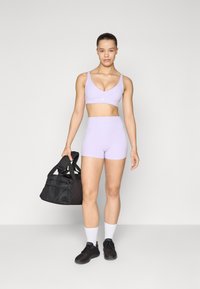 Lys lilla trenings-bh og høyt liv shorts, sammen med svart duffelbagg. Modellen har på seg svarte joggesko og hvite sokker. Glatt stofftekstur.