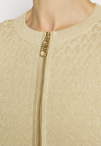 Cardigan con zip color beige, caratterizzato da un sottile motivo testurizzato. Presenta una cerniera dorata e colletto a coste, realizzato in un morbido e leggero tessuto.