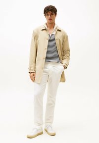 Trench coat beige leggero con bottoni, indossato su un maglione a righe bianco e nero. Abbinato a pantaloni bianchi e sneakers bianche.