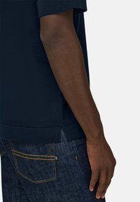 Bras à la peau foncée reposant le long du corps, portant une chemise marine et un jean bleu avec des coutures jaunes et une petite étiquette noire sur la poche.