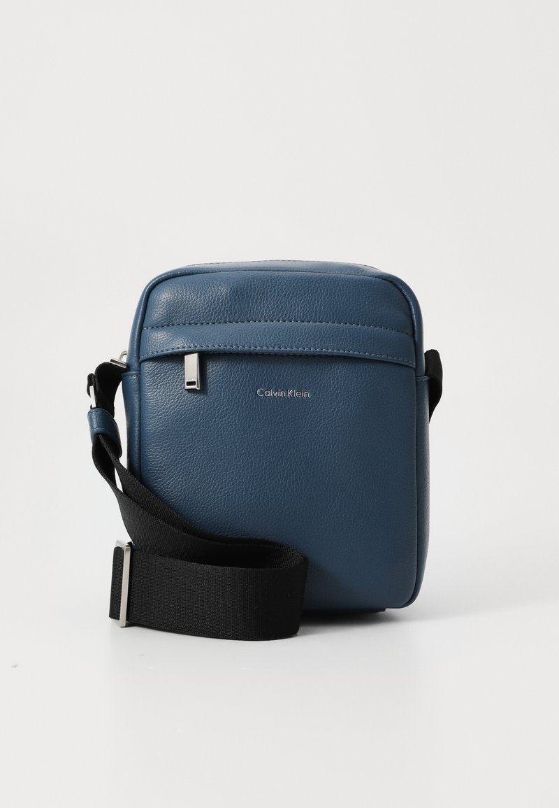 Borsa a tracolla in pelle blu con finitura testurizzata, dotata di una tasca zip frontale, un logo argentato e una tracolla nera regolabile.