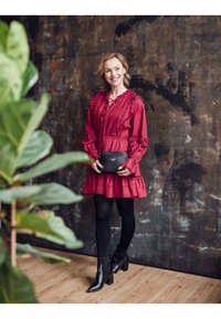 Červené, volánkové šaty s vrstvenou sukní, doplněné černými kotníkovými botami a malou černou kabelkou. Hladká textura, přiléhavý střih. Zelené listy na popředí.