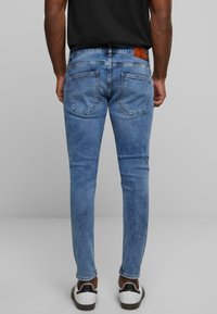 Jeans de mezclilla azul ajustados con un acabado desgastado, diseño de cinco bolsillos y un parche de cuero marrón en la cinturilla. Largo estándar con una textura sutil.